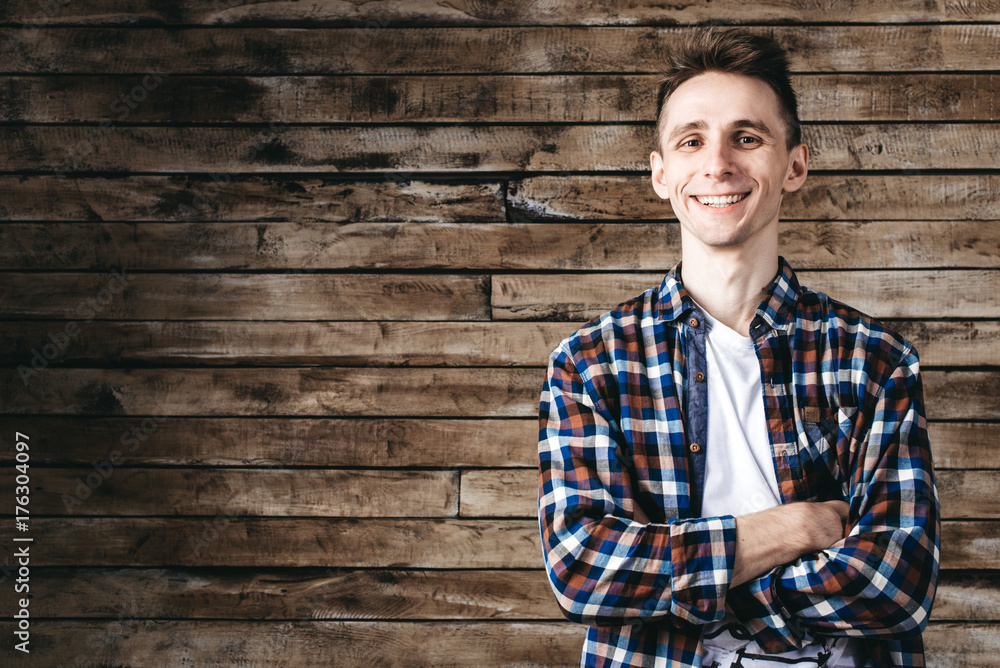 Happy attractive young man in plaid shirt standing and smiling over wooden background Happy attractive young man in plaid shirt standing and smiling over wooden background
