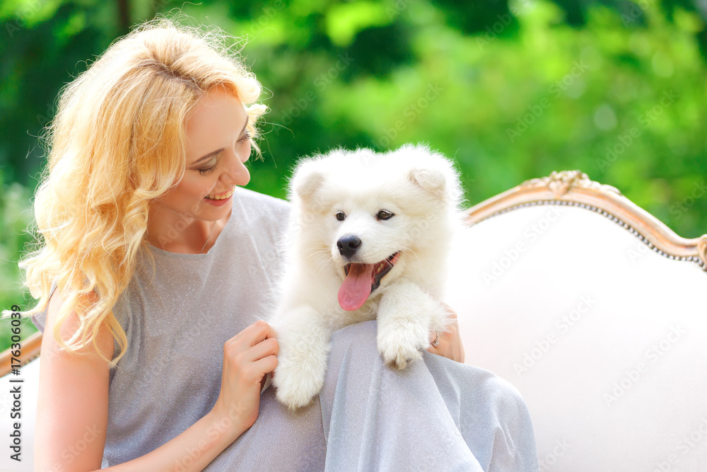 Beautiful young girl with a white puppy in her arms on a retro sofa in a summer garden Beautiful young girl with a white puppy in her arms on a retro sofa in a summer garden