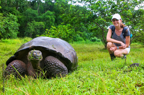 Galapagos giant tortoise with young woman (blurred in background) sitting next to it on Santa Cruz Island in Galapagos National Park, Ecuador