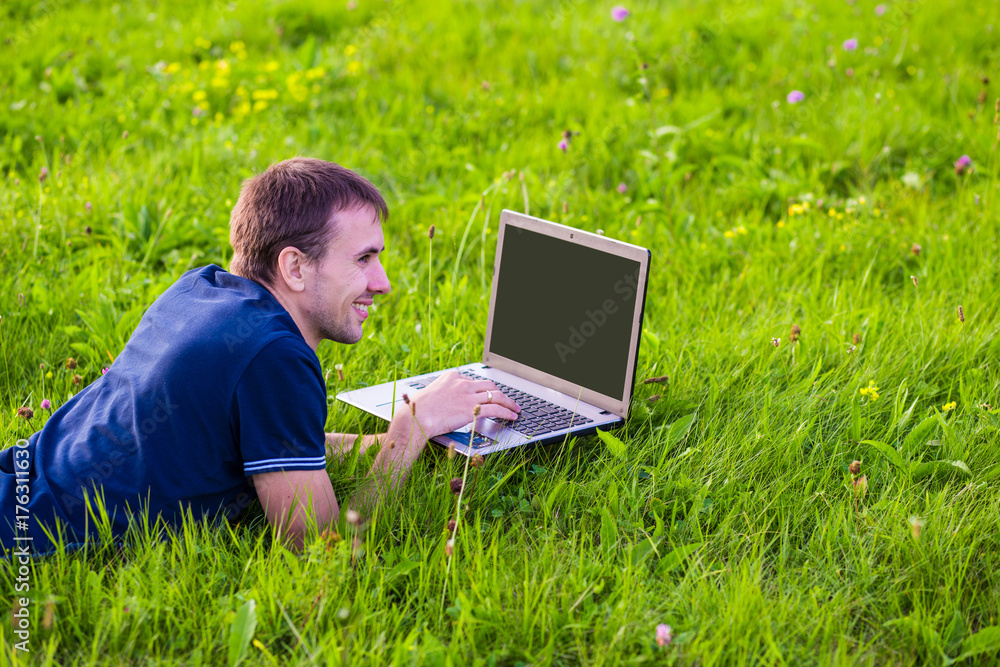Young man lies on green grass at sunset with laptop and working Young man lies on green grass at sunset with laptop and working