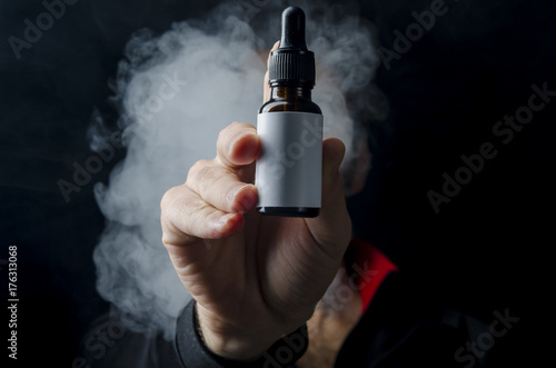 vape e liquid bottle hold by a young man with smoke cloud for electronic cigarette, Isolated eliquid brown bottle for ecig  white empty white label.  photo