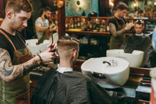 A man getting trendy haircut at barber shop. Male hairstylist in tattoos serving client.