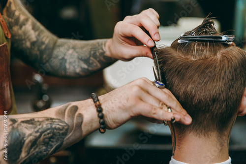 Close up shot of man getting trendy haircut at barber shop. Male hairstylist in tattoos serving client.