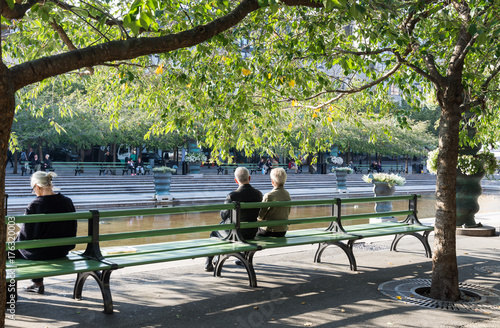 Photography Några personer njuter av solen på bänkarna vid dammen i  Kungsträdgården en fin