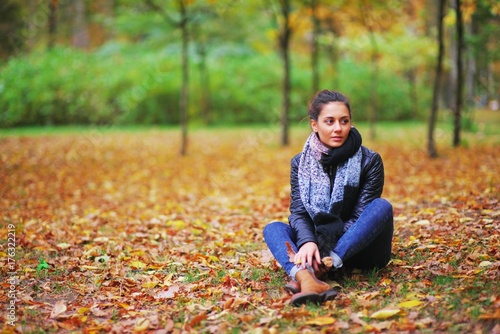 Beautiful black-haired girl in a warm jacket and jeans resting in autumn forest, sitting on the ground.