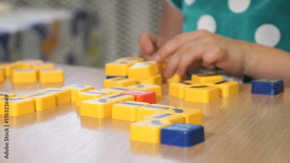 Close-up of unknown little girl in polka dot dress sitting at desk in the kindergarten playing in developing designer and collecting shape in the form of rope on yellow blocks. Close-up of unknown little girl in polka dot dress sitting at desk in the kindergarten playing in developing designer and collecting shape in the form of rope on yellow blocks.