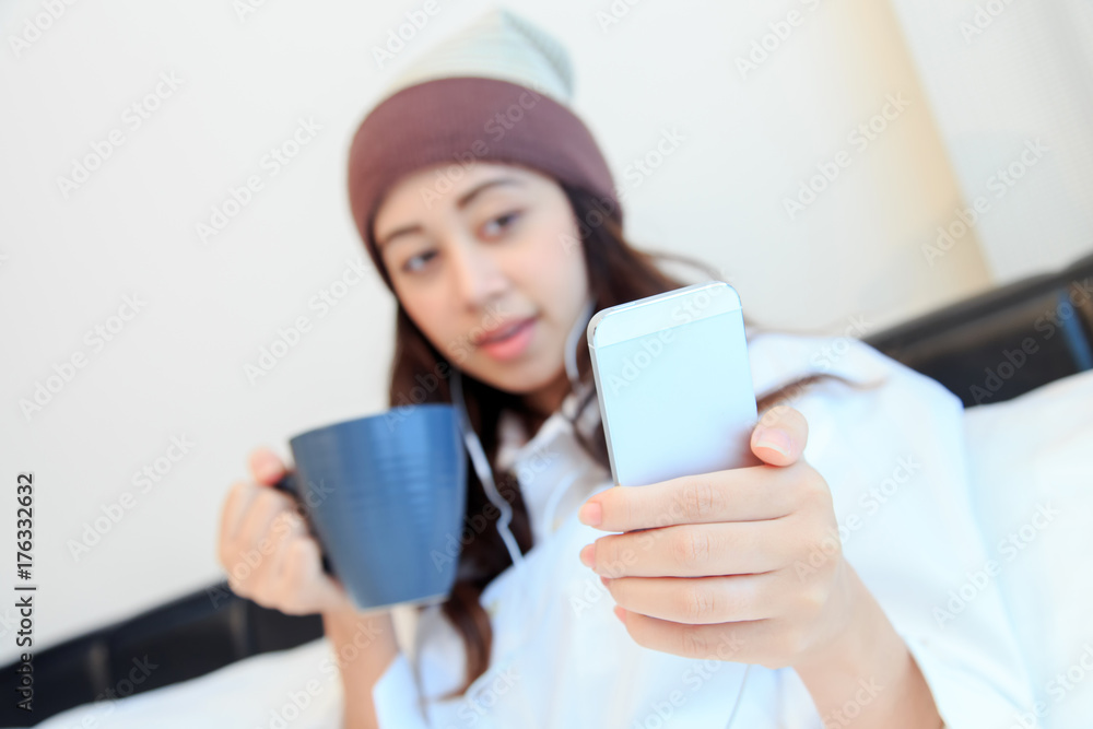 Cheerful young woman sitting in bed texting on her smartphone and drinking coffee at home in bedroom Cheerful young woman sitting in bed texting on her smartphone and drinking coffee at home in bedroom