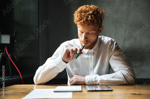 Portrait of thinking young readhead man in white shirt, sitting at wooden table with pen in his mouth