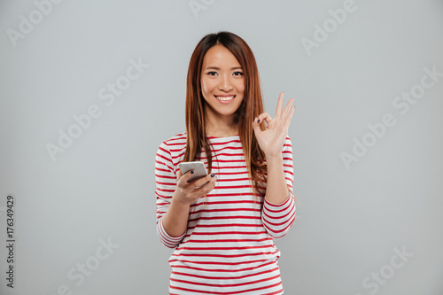 Cheerful young asian woman chatting by phone showing okay gesture.