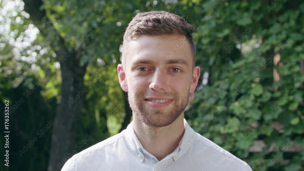 A young man in a white shirt looking at the camera and smiling in the park. Close-up shoot A young man in a white shirt looking at the camera and smiling in the park. Close-up shoot