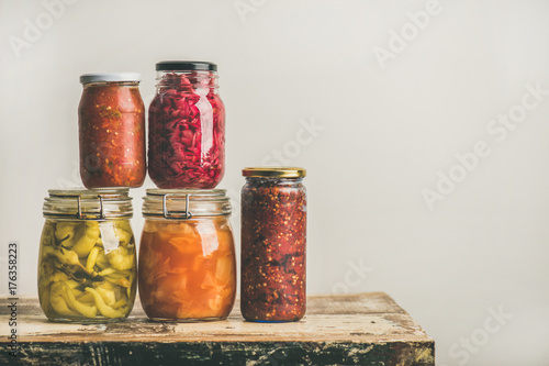 Autumn seasonal pickled or fermented colorful vegetables in glass jars placed in stack over vintage kitchen drawer, white wall background, copy space. Fall home food preserving or canning