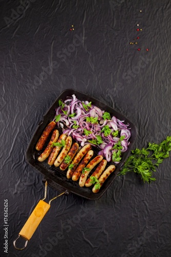 Fried sausages with onion and parsley in a pan on black background