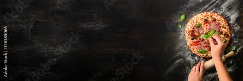 Girl hands making, decopating, preparing pizza with basil leaves on dark background. Top view, copy space. Banner