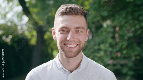 A young man in a white shirt looking at the camera and smiling in the park. Close-up shoot