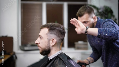 Young barber standing and making stylish haircut of attractive man with clipper in barbershop. Bearded man is sitting on the chair with black protective cape against the mirror and looking at himself.