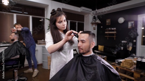 Young female barber making stylish hair to man in salon