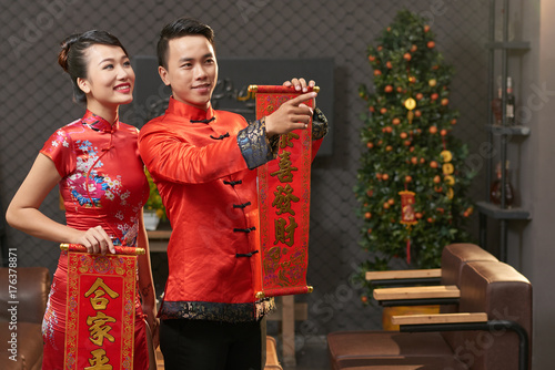 Young Vietnamese couple wearing traditional costumes holding scrolls with New Year couplets in hands while wrapped up in living room decoration