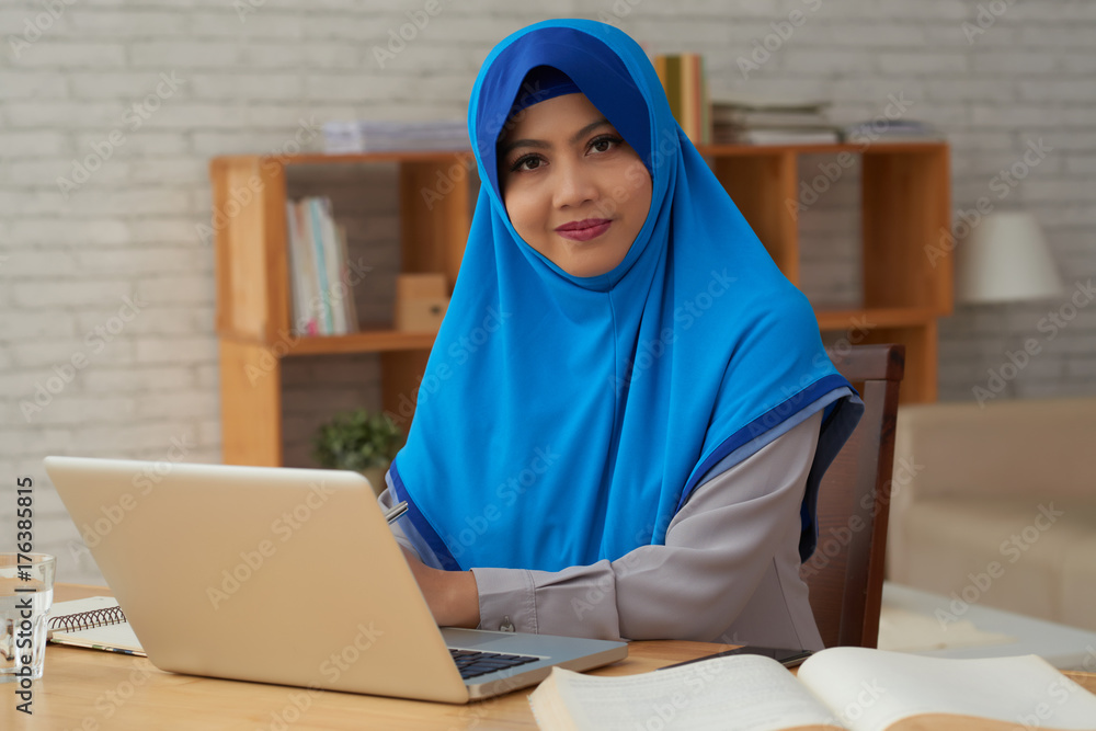 Portrait of attractive woman in hijab sitting at table with notebook and looking at camera Portrait of attractive woman in hijab sitting at table with notebook and looking at camera