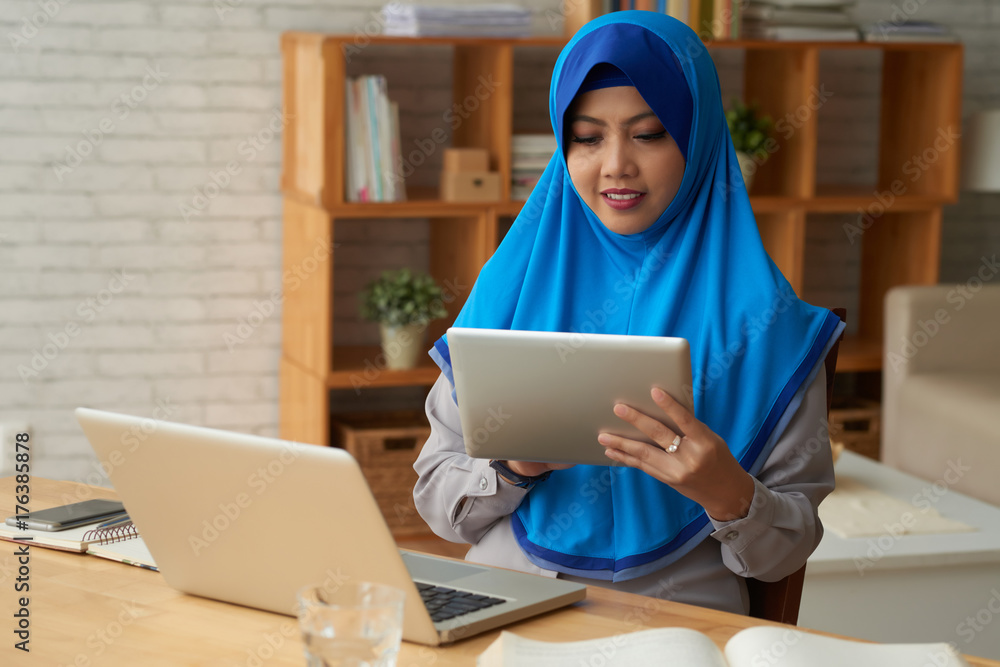 Concentrated young woman in hijab sitting at table with laptop and using digital tablet Concentrated young woman in hijab sitting at table with laptop and using digital tablet