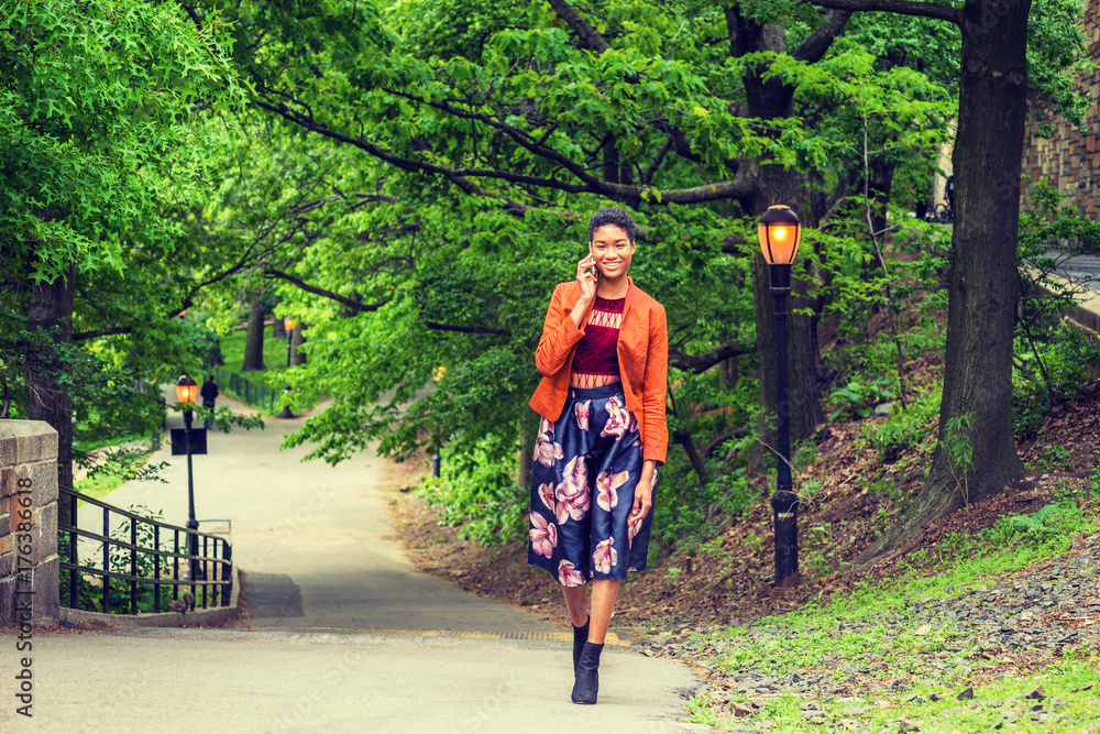 Young African American Woman talking on cell phone outside in New York, wearing orange red jacket, dark purple flower patterned skirt, black boot shoes, walking up on small road with green trees..