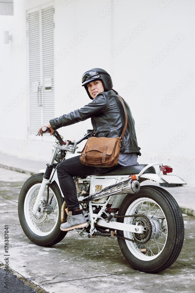 Obraz premium Pełny długość portret ufny brutalny mężczyzna jest ubranym hełm i skórzaną kurtkę pozuje dla fotografii podczas gdy siedzący na rocznika motocyklu przygotowywającym dla szukać przygod