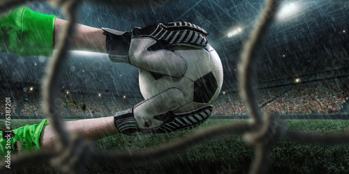 Soccer goalkeeper catches a ball on big professional rainy football arena. View through the football goal. Goalkeeper wears unbranded sport uniform. photo