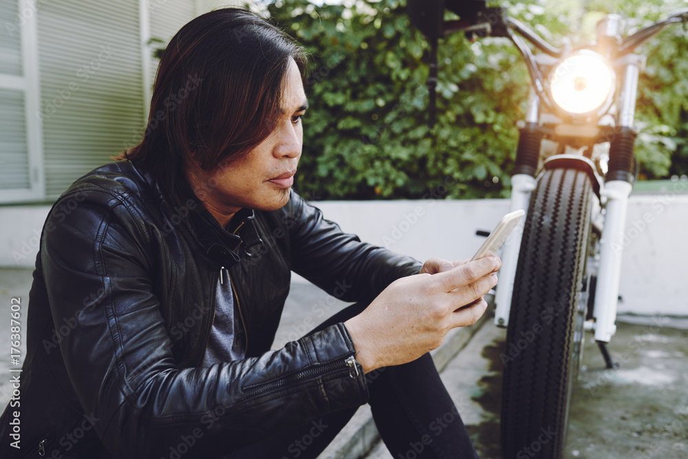 Obraz premium Concentrated Asian biker wearing black leather jacket studying electronic map on smartphone and sitting by side of road while taking break from motorcycle trip, profile view