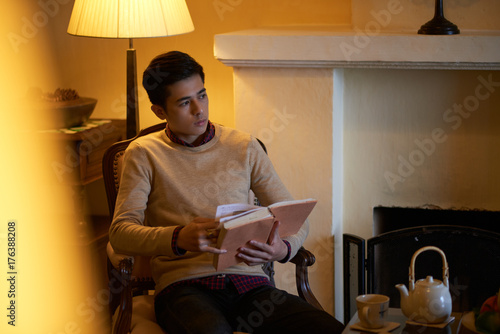 Pensive Asian man sitting in armchair under lamp light with book in his hands