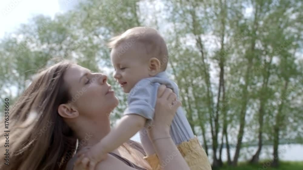 Low angle of beautiful young woman laughing and spinning giggling baby boy, then kissing him on cheek while spending time in park on warm summer day Low angle of beautiful young woman laughing and spinning giggling baby boy, then kissing him on cheek while spending time in park on warm summer day