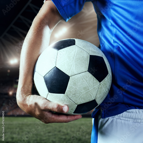 Soccer player holds a football ball on a professional stadium. Player wears blue unbranded uniform. © Alex