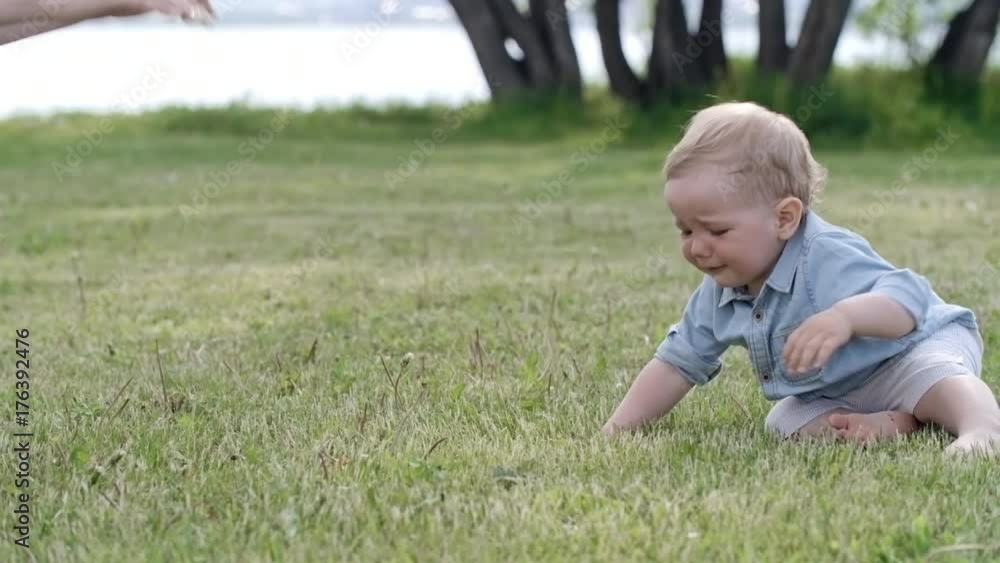 Tracking of adorable toddler boy with blond hair walking barefoot on green grass towards young mother, then falling down and crying Tracking of adorable toddler boy with blond hair walking barefoot on green grass towards young mother, then falling down and crying