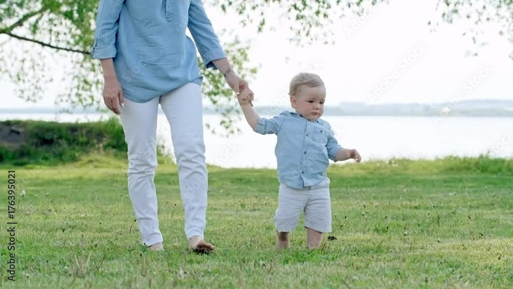 Lockdown of unrecognizable mother holding hand of cute little boy and walking on green grass in park on summer day; lake in background Lockdown of unrecognizable mother holding hand of cute little boy and walking on green grass in park on summer day; lake in background