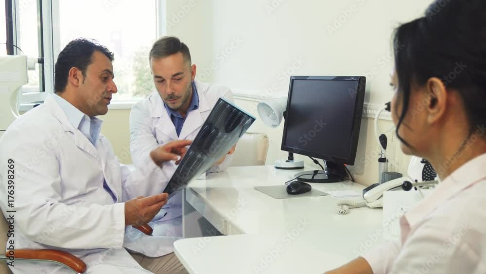 Two doctors and a patient are sitting in the office. One of them has X-Rays in their hands. They examine it and discuss it. The patient is waiting for results Two doctors and a patient are sitting in the office. One of them has X-Rays in their hands. They examine it and discuss it. The patient is waiting for results