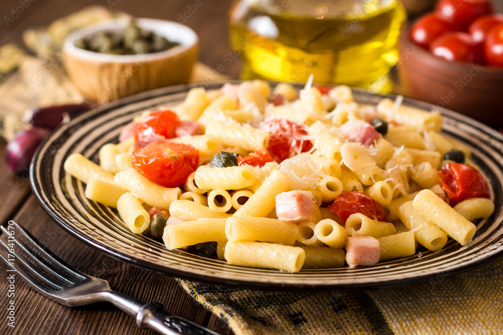 Pasta with bacon, garlic, tomatoes, capers and parmesan cheese in plate on dark wooden background.