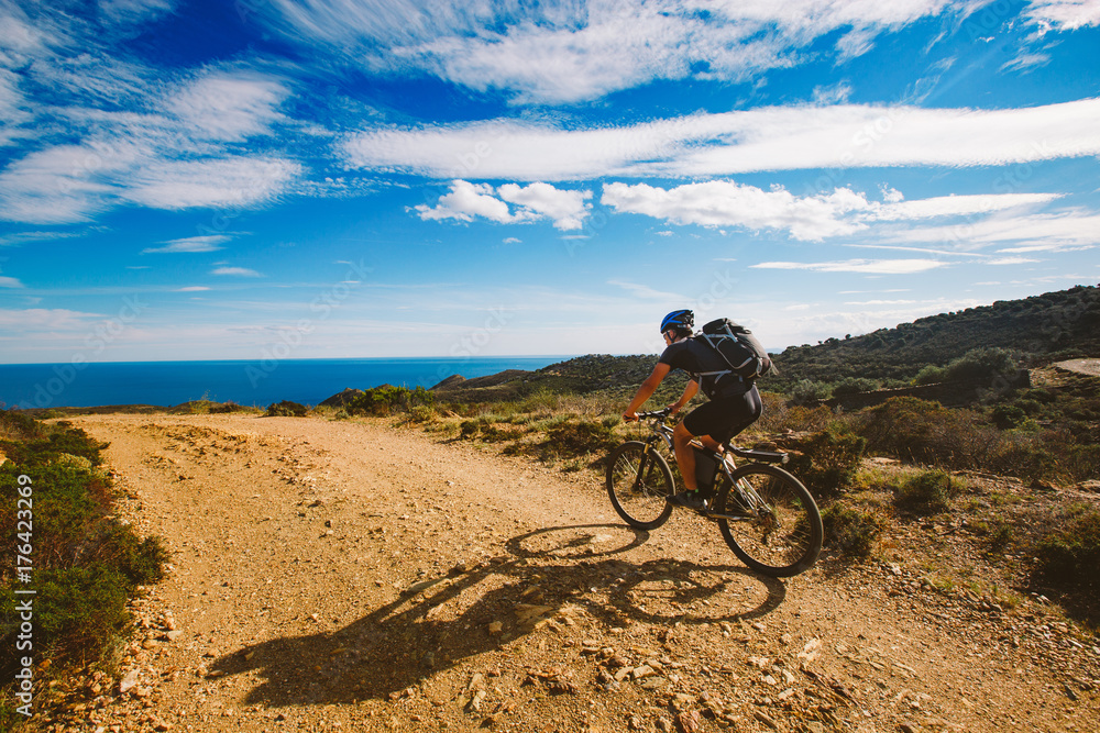 a young guy riding a mountain bike on a bicycle route in Spain on road against the background of the Mediterranean Sea. Dressed in a helmet, a dark one and a black backpack