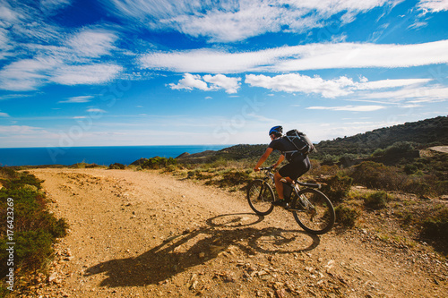Fototapeta Naklejka Na Ścianę i Meble -  a young guy riding a mountain bike on a bicycle route in Spain on road against the background of the Mediterranean Sea. Dressed in a helmet, a dark one and a black backpack