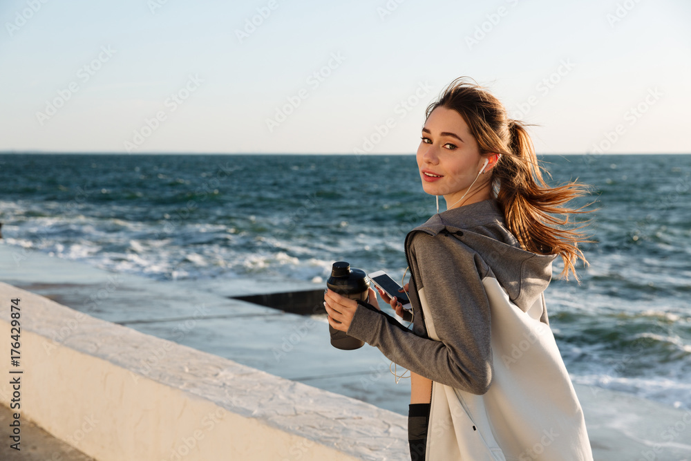 Close-up portrait of pretty smiling sport woman, holding bottle of water while listening to music, seaside outdoor