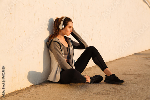 Portrait of cheerful young sport woman listening to music, resting after workout, sitting near white wall © Drobot Dean
