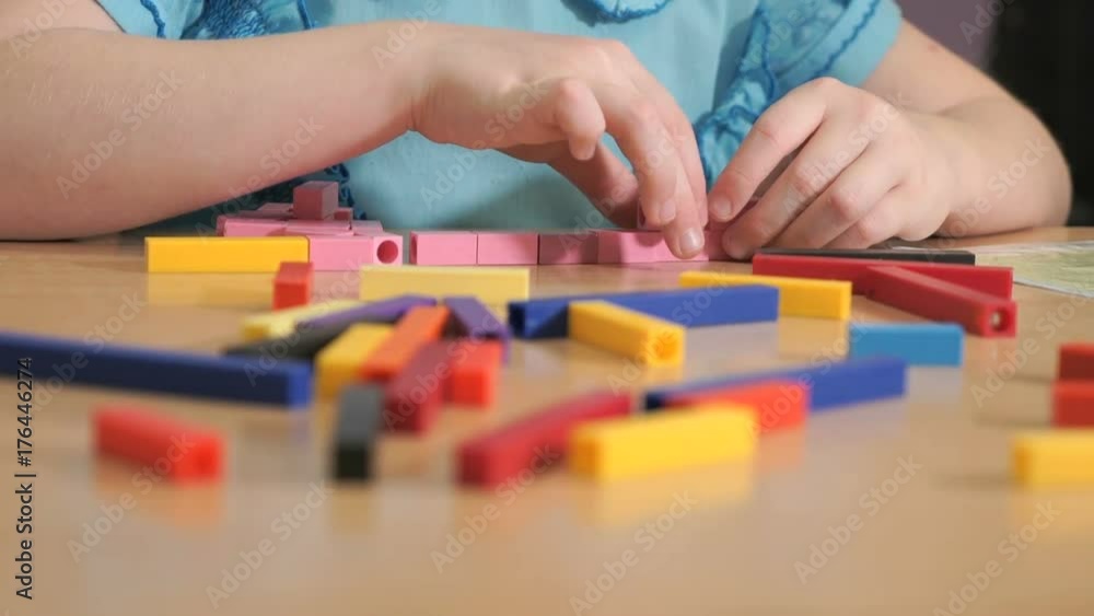 Close-up of hands of unidentified little girl in blue shirt sitting at desk in the kindergarten playing in developing designer - making figures of stripes of various colors. Close-up of hands of unidentified little girl in blue shirt sitting at desk in the kindergarten playing in developing designer - making figures of stripes of various colors.