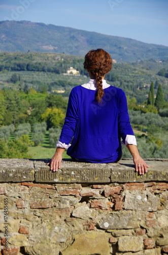 Young woman in blue shirt sitting on a stone wall looking at the Tuscany countryside