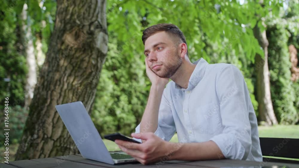 A young man using a phone in the park and sitting at the table. Medium shot. Soft Focus A young man using a phone in the park and sitting at the table. Medium shot. Soft Focus