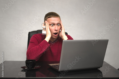 Fotomural A young caucasian man in glasses working at a laptop at the table and in shock from what he saw at computer monitor