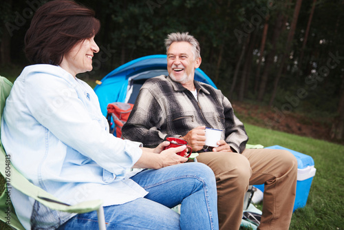 Mature couple sitting in camping chairs, beside tent, holding cups of tea, smiling photo