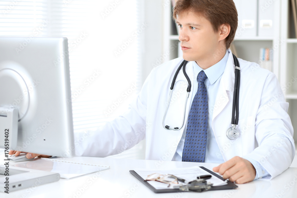 Friendly male doctor is sitting at the table and working in the hospital office. Ready to examine and help patients. High level and quality medical service concept. Friendly male doctor is sitting at the table and working in the hospital office. Ready to examine and help patients. High level and quality medical service concept.