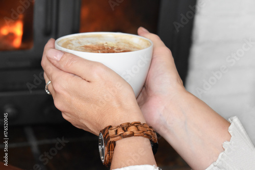 Woman with elegant slender hands holding cappuccino at home in front of cozy burning fire