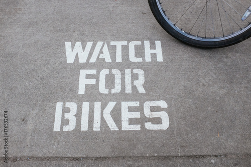 Sign on sidewalk that reads Watch for Bikes with bicycle wheel in corner.