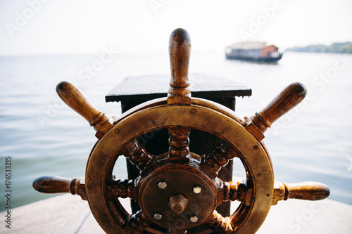 Old boat steering wheel in the sea with boat in the background. Kerala backwaters, India.