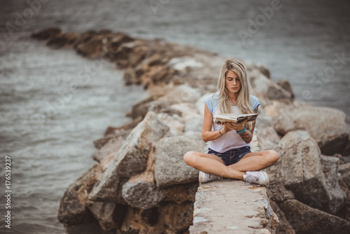 Beautiful blonde woman sitting on a stone embankment and reads book. Hands tattooed. modern, trendy, student