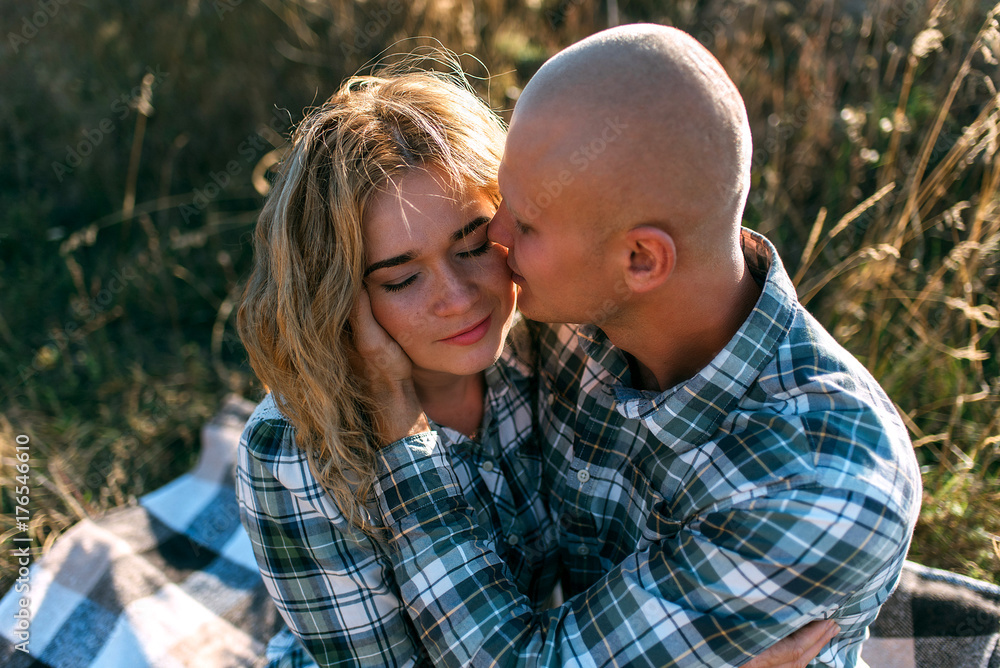 Fototapeta premium man guy in checkered shirt and girl in green dressed in a black shirt and jeans kiss standing in grass sitting in grass half on top of sunset with dog running around fooling around