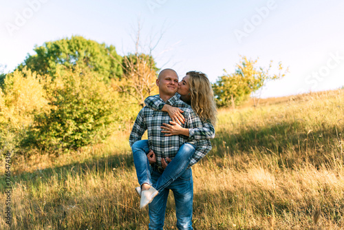 male guy in checkered shirt and girl in green dressed in a black shirt and jeans kiss standing in grass sitting in grass half on top of sunset with dog running around fooling around
